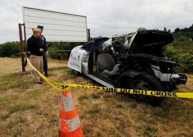 Ein Polizist in Uniform steht neben einem schwer beschädigten Auto an der Straße, mit einem Verkehrskegel und einem Band an einem Pfahl in der Nähe, sowie Bäumen und einem bewölkten Himmel im Hintergrund.