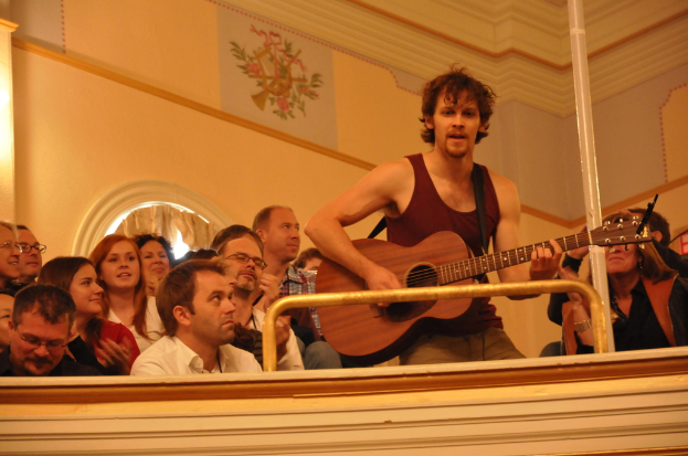Eine Gruppe von Menschen, die Musik hören und genießen, mit einem Mann in der Mitte, der ein ärmelloses Hemd trägt, singt und eine Gitarre spielt, vor einer braunen Wand mit einem Bild oben.