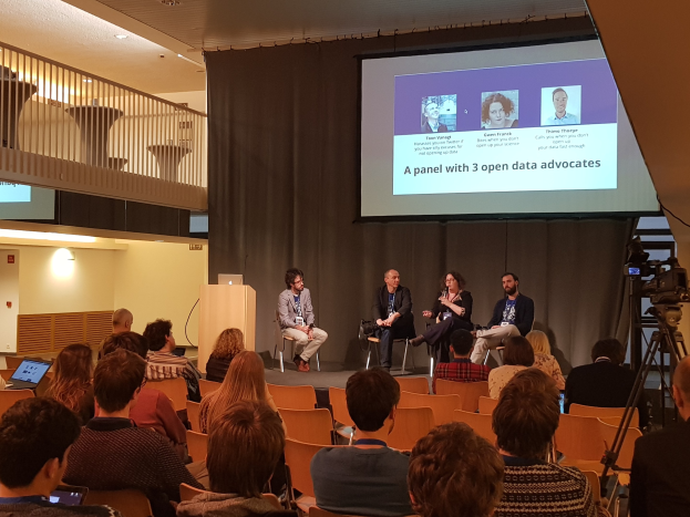 Eine Gruppe von Menschen sitzt auf Stühlen vor einem Projektionsschirm mit einem Laptop auf einem zentralen Tisch, eine Kamera auf einem Ständer rechts daneben und Geländern, Lampen und einer Wand im Hintergrund, die an einer Podiumsdiskussion über Open-Data-Aktivisten teilnehmen.