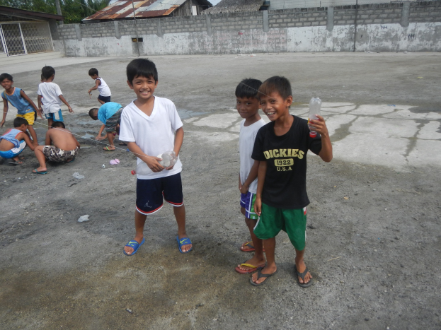 Gruppe junger Jungs, die in einer Parklöcher stehen, einige halten Flaschen, mit Häusern, Bäumen, Masten und einem klaren blauen Himmel im Hintergrund.