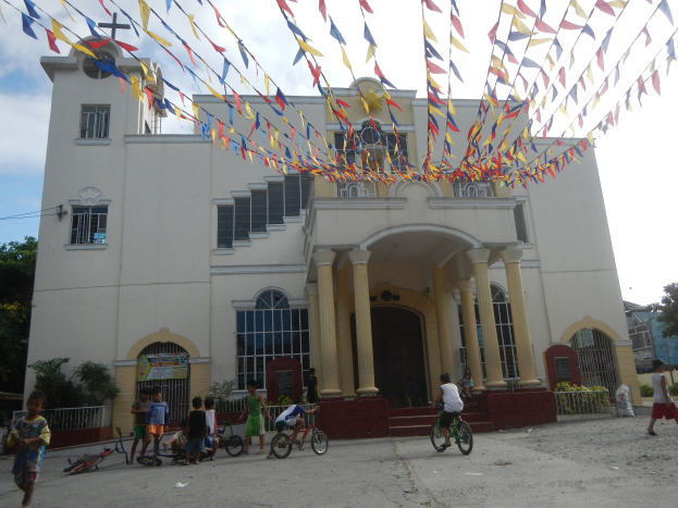 Eine Gruppe von Menschen, die vor einer weißen Kirche mit architectural details Fahrräder fahren, umgeben von Grün, einem Banner, Bäumen und einem bewölkten Himmel, mit bunten Fahnen, die an einem Seil befestigt sind.