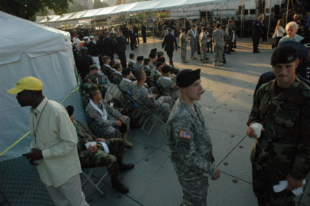 Eine Gruppe von Menschen in Militäruniformen versammelt sich im Freien mit Zelten und einem Baum im Hintergrund, einige sitzen auf Stühlen in der Mitte.