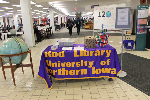 Eine Ausstellung mit einem Tisch, auf dem verschiedene Landesflaggen, eine Box, Papiere und ein Banner stehen, eine Kugel links daneben, eine Anschlagtafel rechts daneben und mehrere Tische und Stühle in der Ferne.
