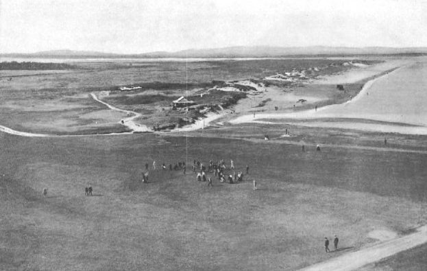 Schwarzes und weißes Foto von Golfern auf dem 18. Loch auf dem Royal Birkham Golf Club, mit saftig grünem Rasen, verstreuten Häusern, sanften Hügeln und einem hellblauen Himmel im Hintergrund.