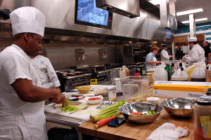 Eine Gruppe von Köchen in einer Küche bei der Zubereitung von Speisen auf einem Schneidbrett, mit einem Koch im Vordergrund in einer weißen Schürze und Mütze, der ein Messer hält und von verschiedenen Utensilien, Schüsseln, Gemüse, einem Mobiltelefon, Flaschen und anderen Gegenständen umgeben ist, mit einem Fernseher und Deckenleuchten im Hintergrund.