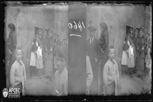 Ein Schwarz-Weiß-Foto einer Gruppe von Menschen, die vor einem Gebäude mit einer Wand im Hintergrund stehen, mit Text, der darauf hinweist, dass es sich um Flüchtlinge handelt, und einem Logo mit der Aufschrift "Aktusi fotografie oil center éxigronostia de catalunya" in der unteren linken Ecke.