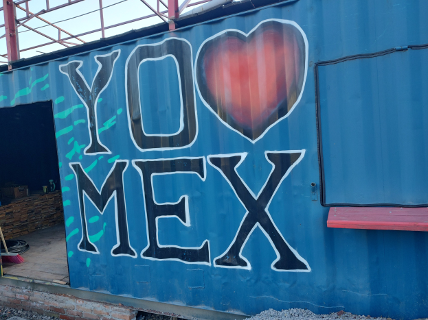Blauer Seecontainer mit "Ich liebe Mexiko" darauf, umgeben von Steinen unten und einem Schuppen im Hintergrund.