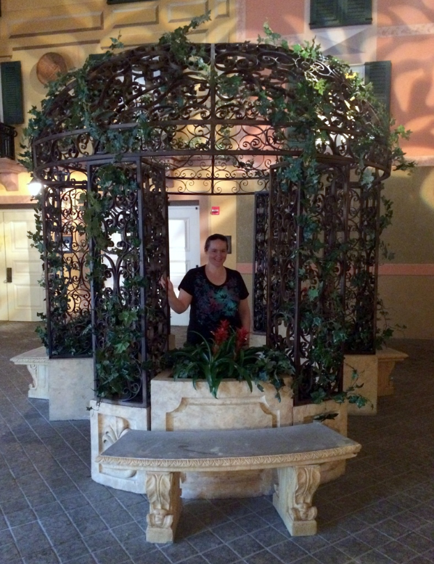 Eine Bank im Vordergrund mit einer verzierten Eisen Säule und Kletterpflanzen dahinter, die eine Frau in einem schwarzen Kleid und ein Gebäude mit Wänden, Fenstern und Türen im Hintergrund zeigt.