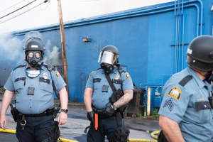 Drei Polizisten in Einsatzausrüstung, einschließlich Helmen und Gasmasken, gehen eine Straße mit einer blauen Wand, Rohren und einem Strommast mit Drähten im Hintergrund hinunter, unter einem sichtbaren Himmel.