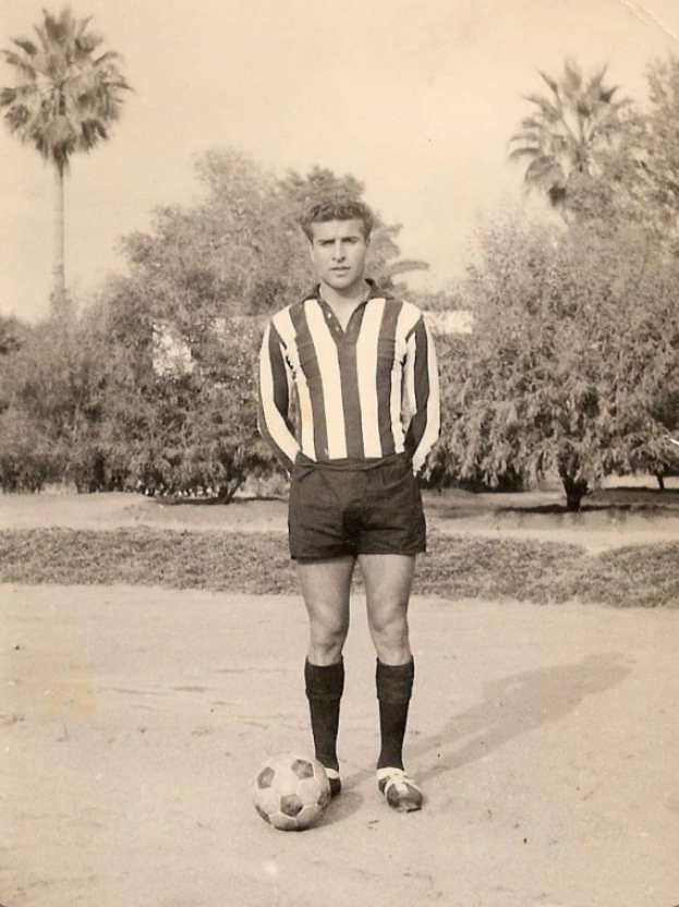 Ein schwarz-weisses Foto eines alten Mannes, der einen Fussball halt, mit Baumen und Himmel im Hintergrund.
