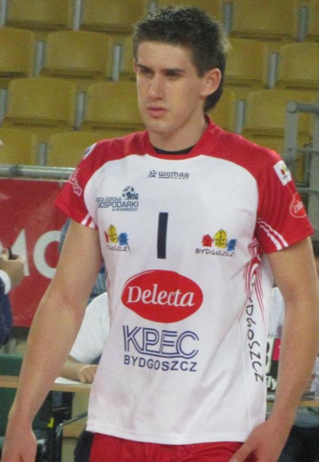 Ein Mann in rot-weißer Handballdressur steht auf einem Feld, umgeben von Zuschauern, mit einem roten Banner im Hintergrund, das 'Krasnoyarsk vs Bydgoszcz' zeigt und entschlossen aussieht.