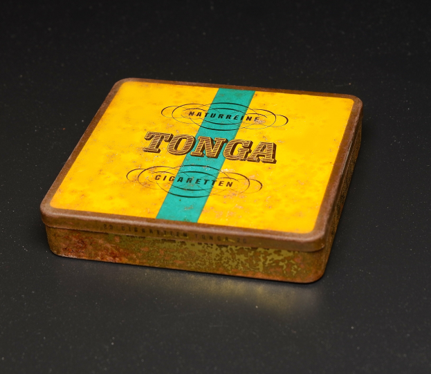 Ein gelb-blauer Blechbehälter mit Tonga Zigaretten auf einem schwarzen Tisch, mit Text, der die Marke und Art der Zigaretten angibt.