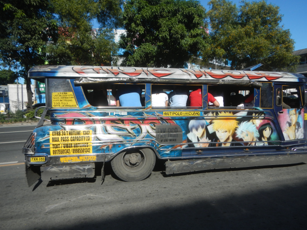 Ein blauer Bus mit Anime-Charakteren, die auf der Seite gemalt sind, fährt eine Straße entlang, die von Bäumen, Gebäuden und einem klaren blauen Himmel umgeben ist, mit ein paar Menschen drinnen sichtbar.