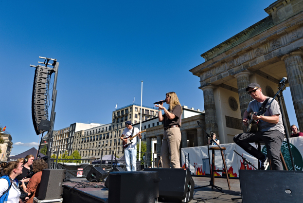 Eine Gruppe von Menschen, die auf einer Bühne Musik spielen, mit dem Brandenburger Tor im Hintergrund, begleitet von Lautsprechern und anderem Equipment unter einem klaren Himmel.