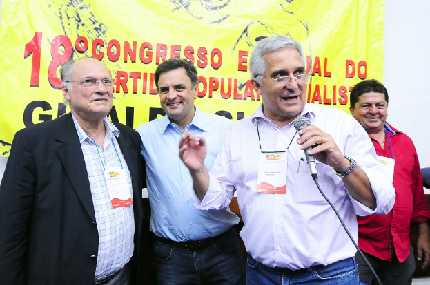 Ein Mann spricht in ein Mikrofon vor einem gelben Vorhang mit "18 Grad Kongress O" darauf, während andere Männer im Hintergrund stehen.