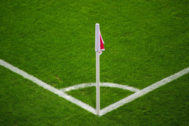 Ein Fußballfeld mit grünem Rasen, weißen Begrenzungslinien und einem Elfmeterpunkt, der durch einen weißen Pfosten mit einer roten und weißen Fahne markiert ist.