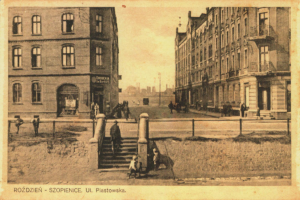 Ein altes Foto einer Stadtstraße mit Gebäuden, Menschen, Treppen, Pfählen und einem Zaun, mit Text unten.