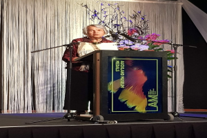 Eine Frau steht an einem Podium mit Mikrofon, Blumen und einer Texttafel vor einem Vorhang und einer Wand bei der jährlichen Verleihung des Auckland Museum of Art Awards.