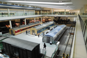Ein großer Museumsraum mit verschiedenen Bahnen in unterschiedlichen Größen und Farben, mit Menschen, die die Ausstellungsstücke beobachten und Deckenlampen, die den Raum beleuchten.