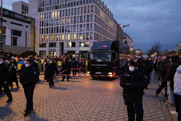 Eine Gruppe von Menschen steht vor einem Lkw auf einer Straße, umgeben von Gebäuden, Laternen, Bäumen und einem bewölkten Himmel, wobei einige eine Mütze und eine Maske tragen und ein Band mit einem Pfahl im Vordergrund zu sehen ist.