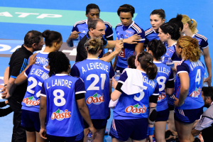 Eine Gruppe von Frauen-Handballspielerinnen sitzt zusammen auf einem Platz, einige halten Flaschen, während jemand auf der rechten Seite mit einer Kamera das Geschehen einfängt.