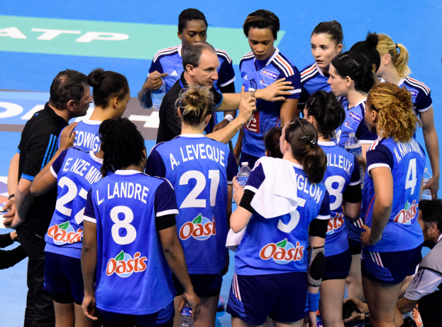 Eine Gruppe von Frauen-Handballspielerinnen sitzt zusammen auf einem Platz, einige halten Flaschen, während jemand auf der rechten Seite mit einer Kamera das Geschehen einfängt.