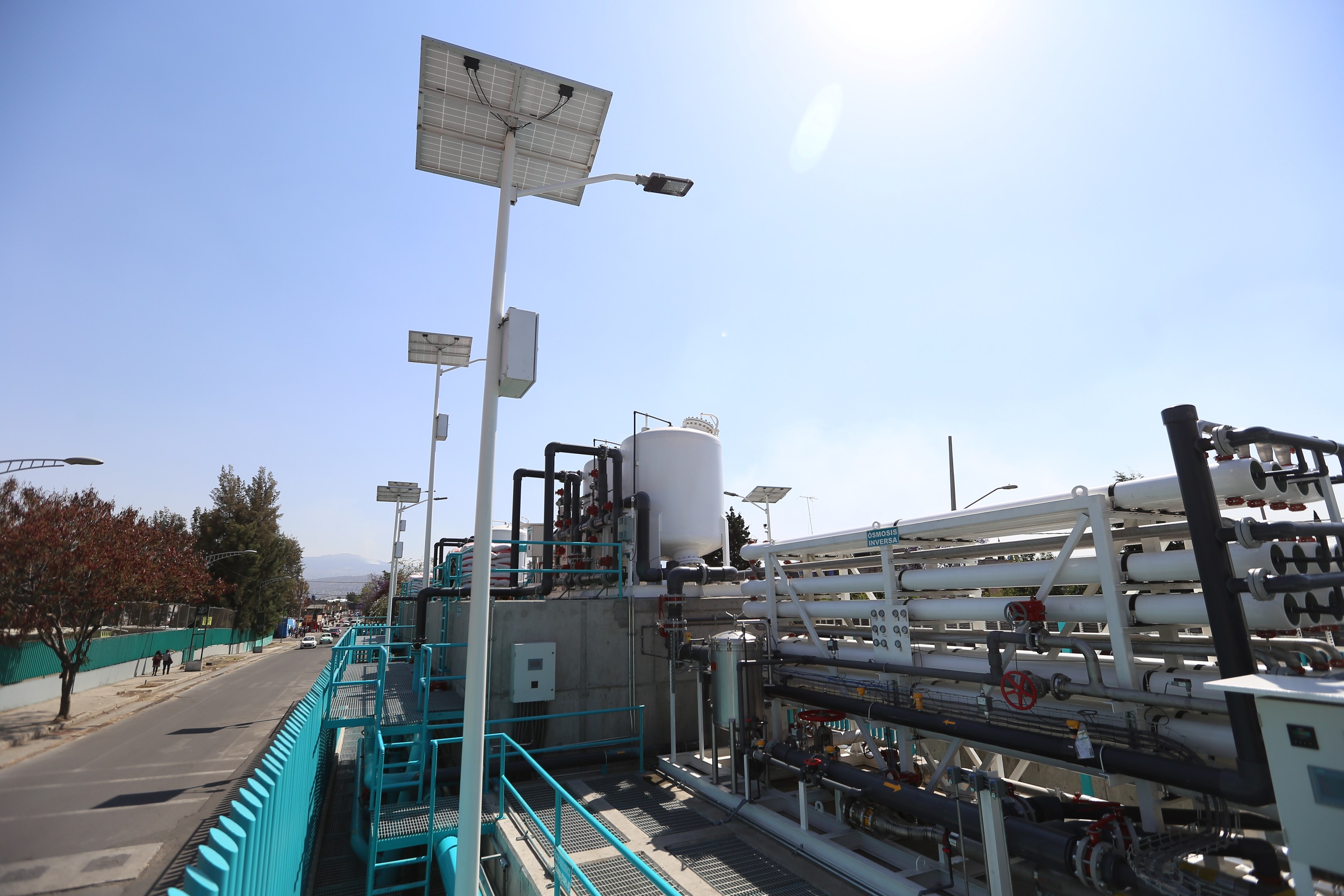 Großes industrielles Gebäude mit ausgedehnten Rohrleitungen und Solarpaneelen auf dem Dach, umgeben von Geländern, Masten, Laternen, Schildern, Bäumen und Fahrzeugen auf der Straße, gelegen in der Nähe von Hügeln und einem klaren blauen Himmel, Teil der neuen Wasseraufbereitungsanlage des California Department of Environmental Protection