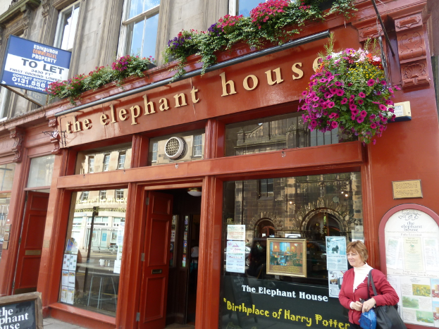 Ein Glasfenster mit einem "Zu-Vermieten"-Schild und einer Nummer darüber, ein Elefantenhaus mit Blumenpflanzen, eine Frau mit einer schwarzen Handtasche, die eine blaue Plastikhülle hält, eine Eingangstür, eine weitere Tür links und ein Tisch drinnen mit einem "Elefantenhaus"-Schild.