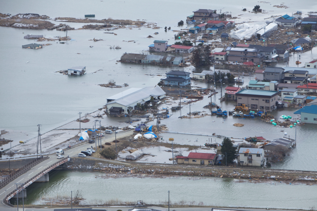 Eine Luftaufnahme einer überfluteten Stadtlandschaft mit versunkenen Gebäuden, Bäumen, Polen, Fahrzeugen und einer Brücke, die die Verwüstung durch das Japan-Erdbeben und den Tsunami zeigt.