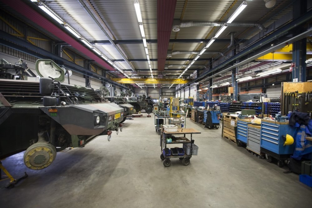 Großes Lager mit Maschinen, Fahrzeugen links, Wagen mit Gegenständen rechts und Deckenbeleuchtung.