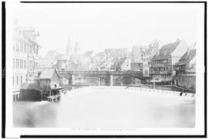 Ein Schwarz-Weiß-Foto eines Flusses mit einer Brücke darüber, umgeben von Gebäuden mit Fenstern, Bäumen und einem Himmel im Hintergrund, mit dem Text "Nürnberg, Blick von Karl's Brücke" unten.
