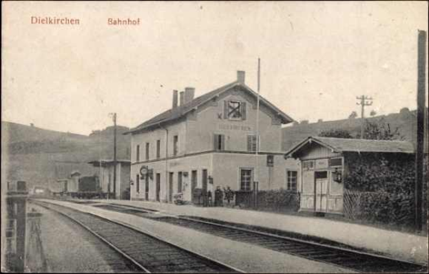 Schwarzes und weißes Foto einer Bahnstation in Dielkirchen, Deutschland, mit einem Zug auf den Schienen, umgeben von Häusern, Pfählen, Bäumen, Pflanzen und Menschen, Hügel im Hintergrund und einem klaren Himmel.