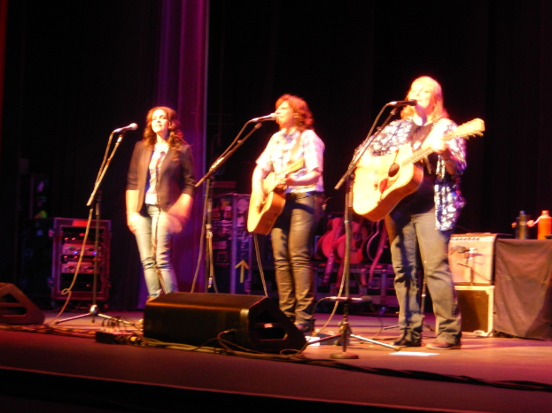 Drei Frauen performen auf der Bühne eines musikalischen Konzerts, jede singt in ein Mikrofon und spielt eine Gitarre, mit einem Tisch voller Wasserflaschen und Gitarren dahinter.