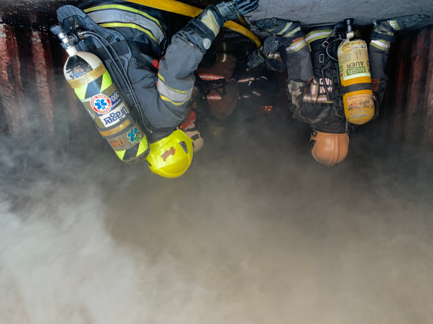 Eine Gruppe von Feuerwehrmännern in Helmen, Handschuhen und Sauerstoffzylindern arbeitet daran, ein Feuer in einem Tunnel zu löschen, wobei Rauch aus der Unterseite aufsteigt.