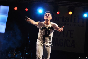 Ein Mann auf einer hell erleuchteten Bühne mit einem Mikrofon in der Hand, im Hintergrund verschiedene Musikinstrumente und ein "Nouveau Magica"-Plakat.