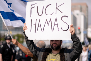 Ein Mann hält ein Schild mit der Aufschrift "Fuck Hamas" vor einer Menge, einige tragen Helme und halten Fahnen, mit Gebäuden und einem klaren blauen Himmel im Hintergrund.