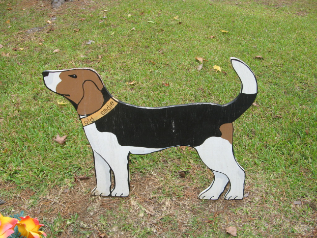 Ein hölzernes Brett mit einer Hundendarstellung liegt auf einer Oberfläche mit Erde, Gras und ein paar Blumen in der linken unteren Ecke.