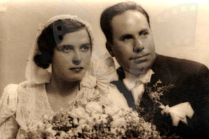 Ein Mann und eine Frau in festlicher Kleidung stehen nebeneinander und strahlen glücklich auf ihrem Hochzeitsfoto, wobei die Frau einen Blumenstrauß hält.