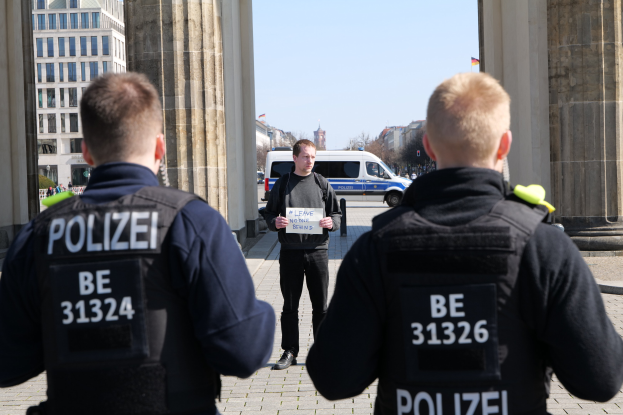 Drei Polizeibeamte stehen vor einem Gebäude, ein Mann in der Mitte hält ein Papier, flankiert von Säulen, während Fahrzeuge, Menschen, Bäume und Gebäude den Hintergrund unter dem Himmel füllen.